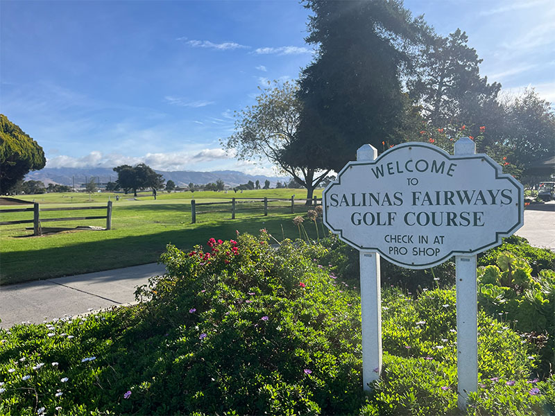 Welcome sign for Salinas Fairways Golf Course beside the course with fairways and mountains in the background.